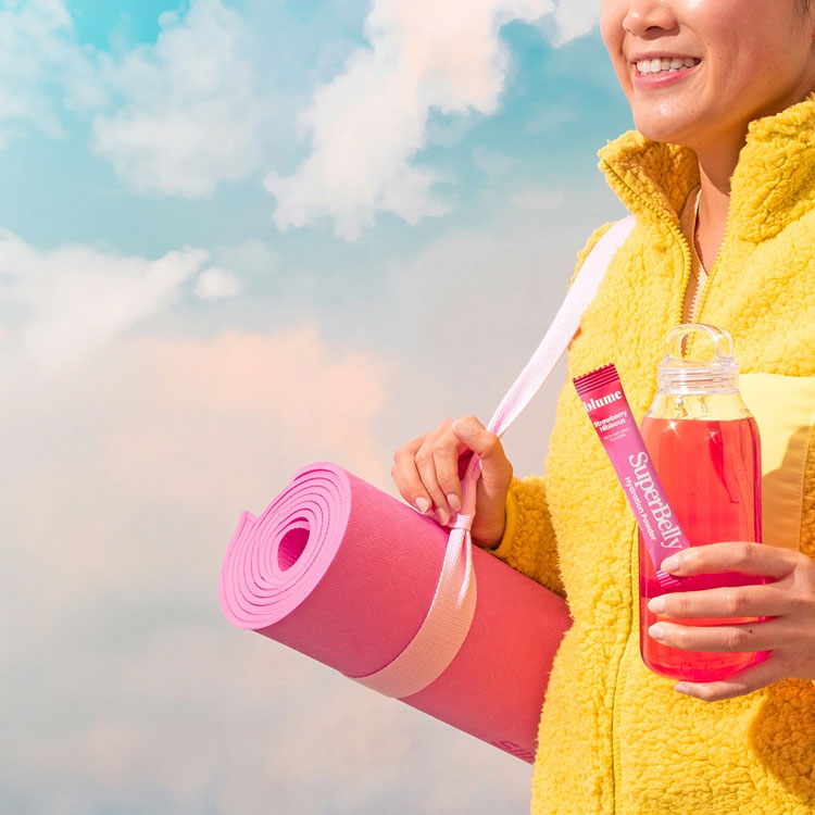 Smiling person in a yellow fleece jacket holds a pink yoga mat and a bottle of pink drink against a cloudy sky background.